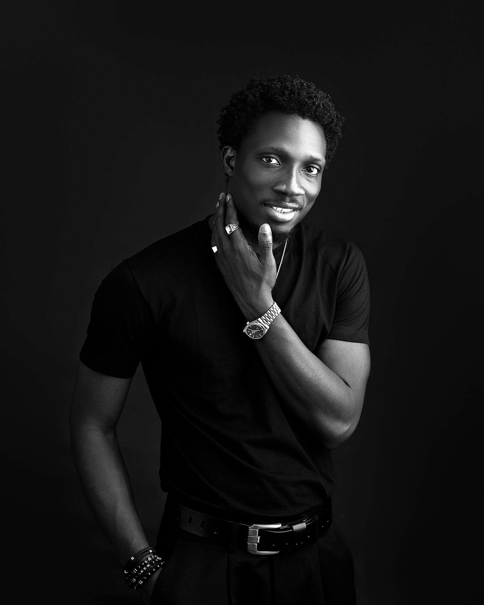 Greatman Takit standing in a black T-shirt, one hand raised to his jaw.