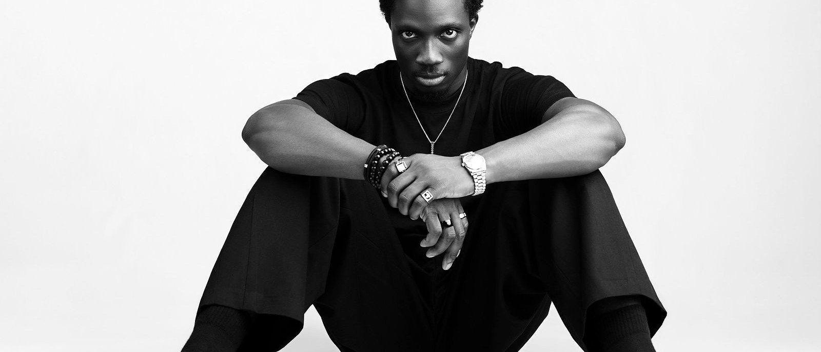 Wide frame of Greatman Takit seated on the studio floor, looking to camera.