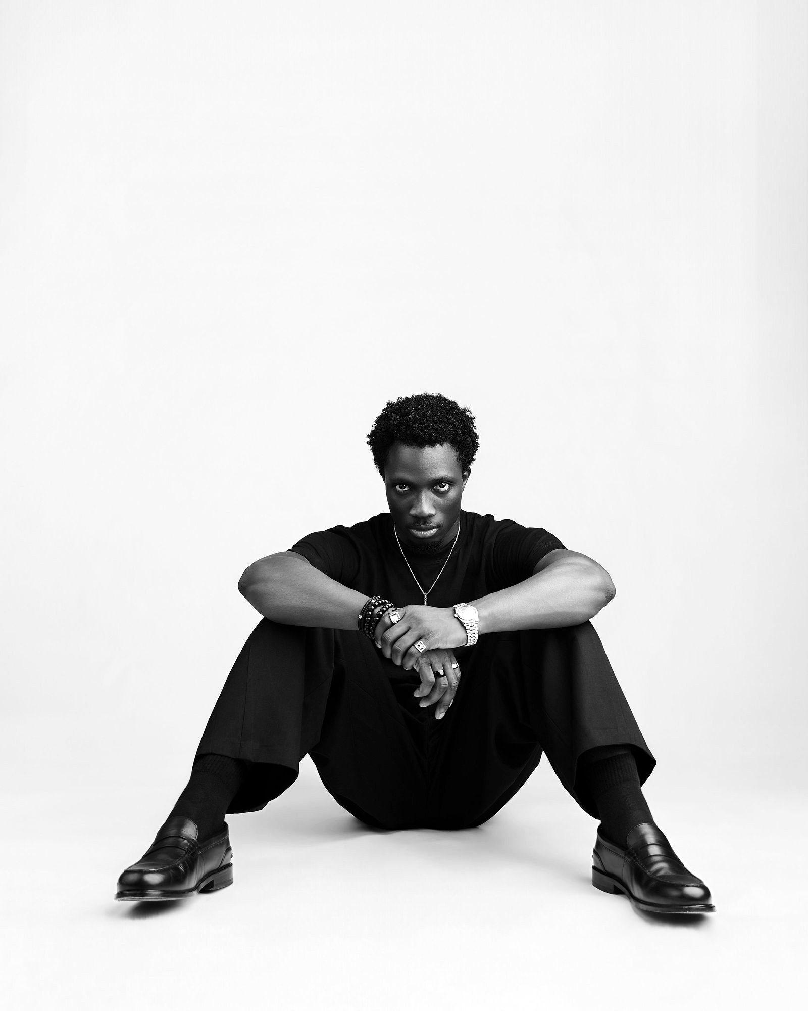 Greatman Takit sitting on the floor of a bare studio, dressed in black.