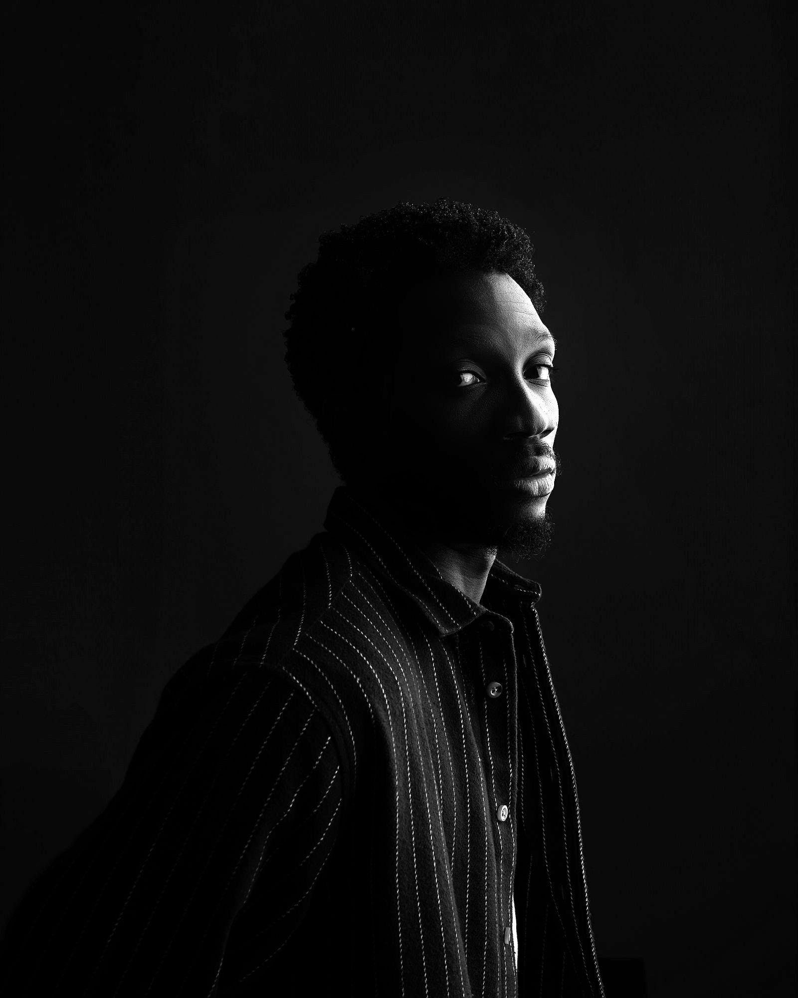 Greatman Takit in profile, half his face lit against a black background.